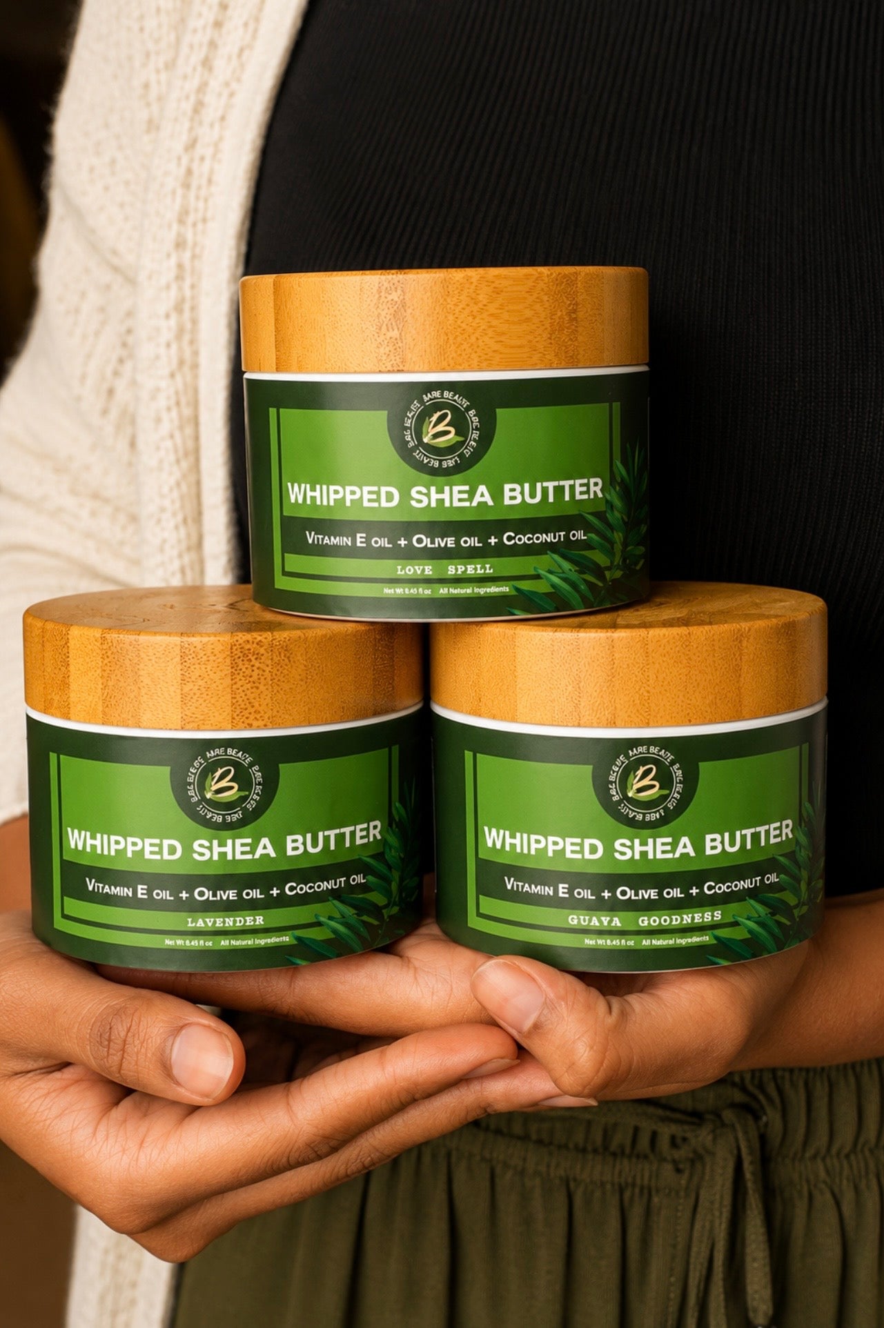 Three jars of Whipped Shea Butter stacked in a pyramid, held in hands against a person's black top and cream cardigan. The jars have light wood lids and dark green labels with white text.