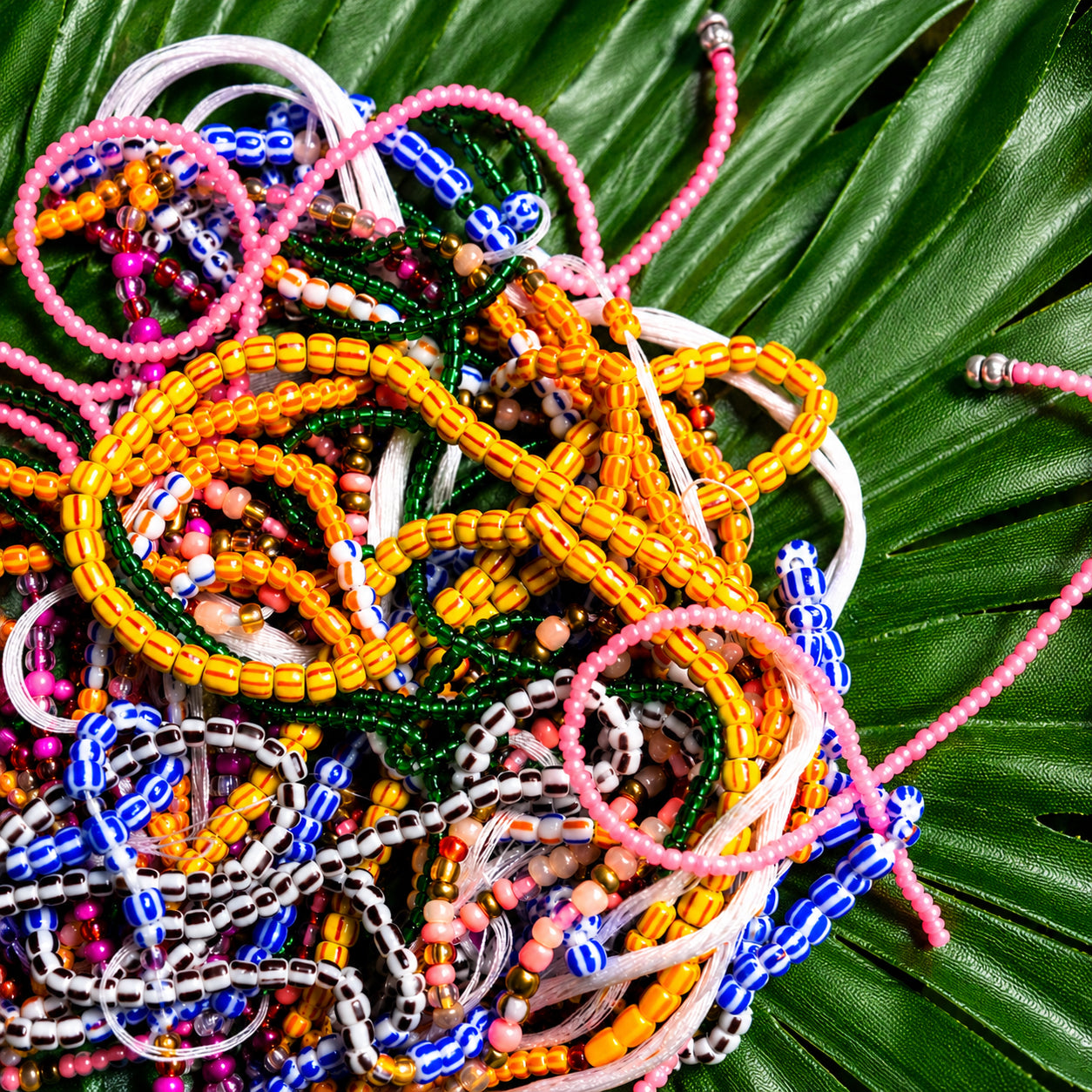 A pile of colorful beaded bracelets and necklaces arranged on a large green palm leaf. The jewelry features multiple bead colors including pink, yellow, blue, green, white, and brown.