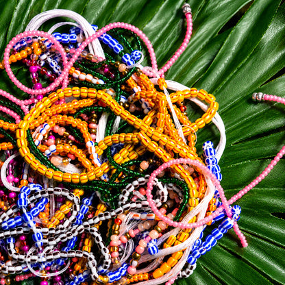 A pile of colorful beaded bracelets and necklaces arranged on a large green palm leaf. The jewelry features multiple bead colors including pink, yellow, blue, green, white, and brown.