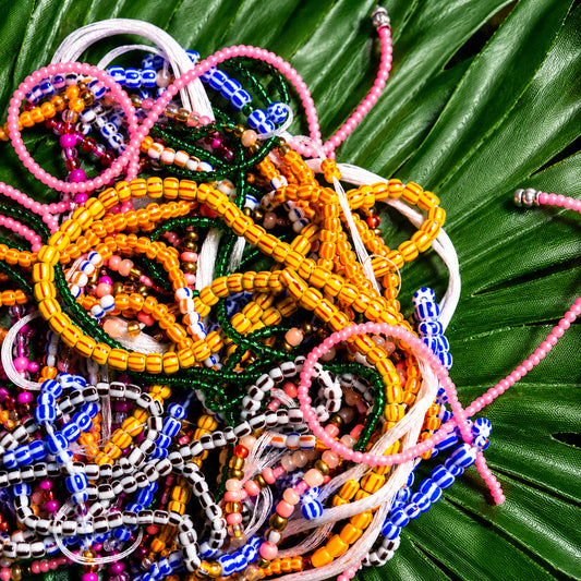 A pile of colorful beaded bracelets and necklaces arranged on a large green palm leaf. The jewelry features multiple bead colors including pink, yellow, blue, green, white, and brown.