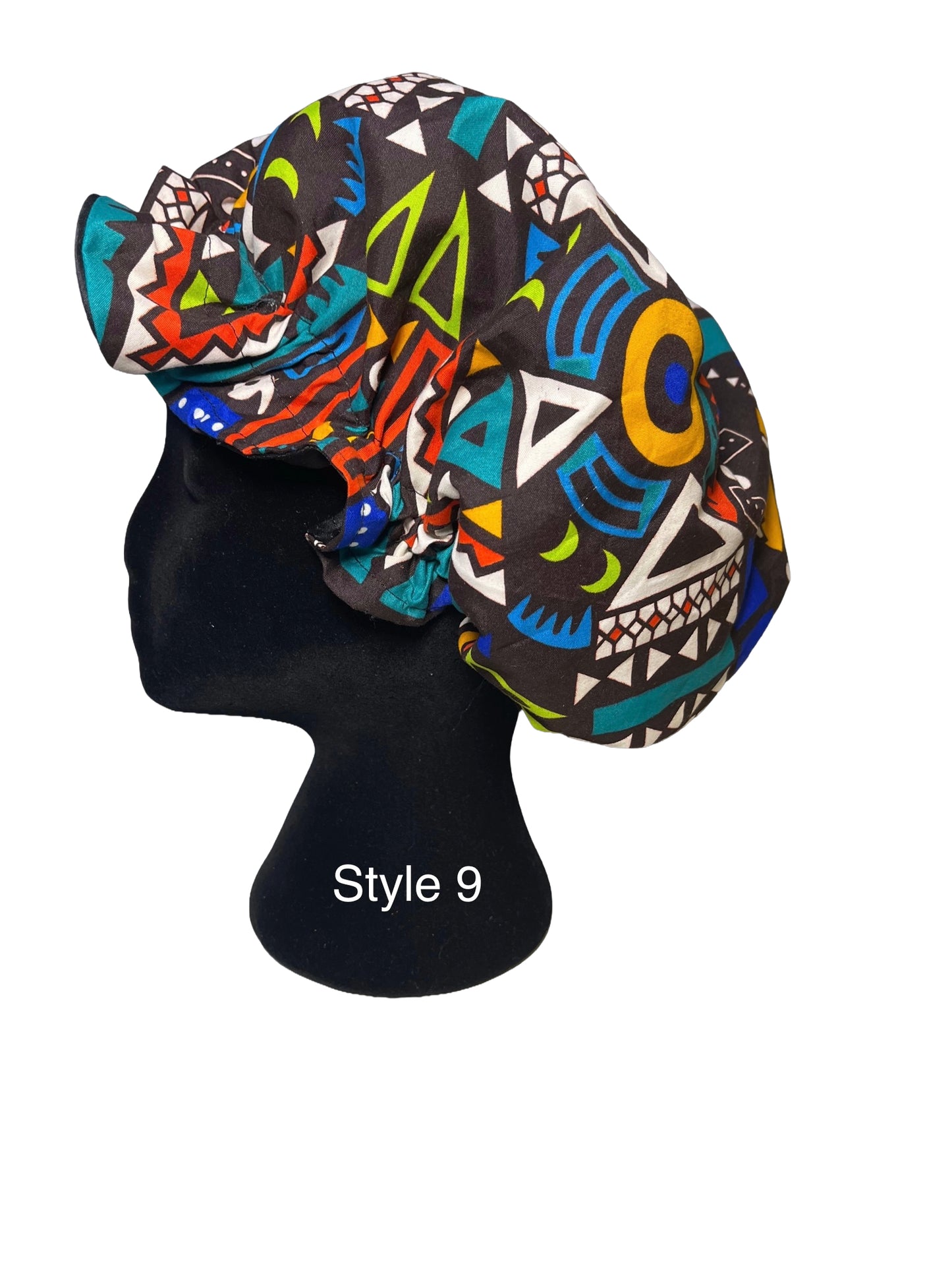 A multicolor African wax print bonnet with geometric patterns, displayed on a black mannequin head. The pattern features black, teal, orange, yellow, green, and white shapes.