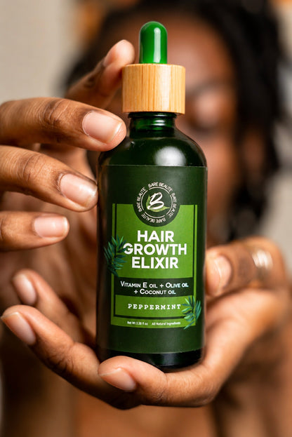 A person holds a dark green glass dropper bottle labeled 'Hair Growth Elixir' with a light wood cap and green dropper top. The background is a softly blurred warm brown/beige setting.