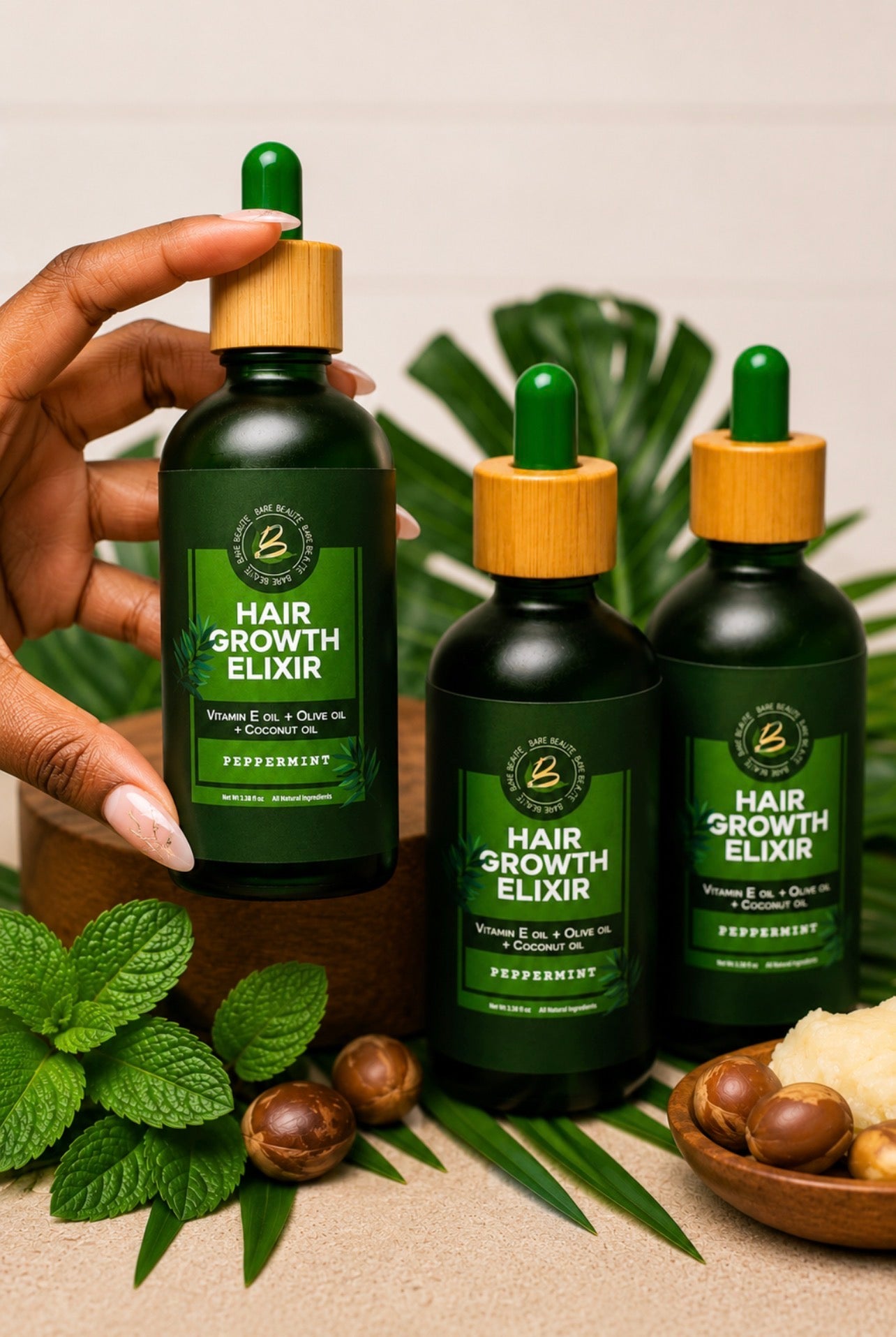 Three dark green bottles of Hair Growth Elixir with bamboo droppers are arranged on a beige surface with mint leaves, macadamia nuts, and a hand holding one bottle. The background features blurred green leaves.