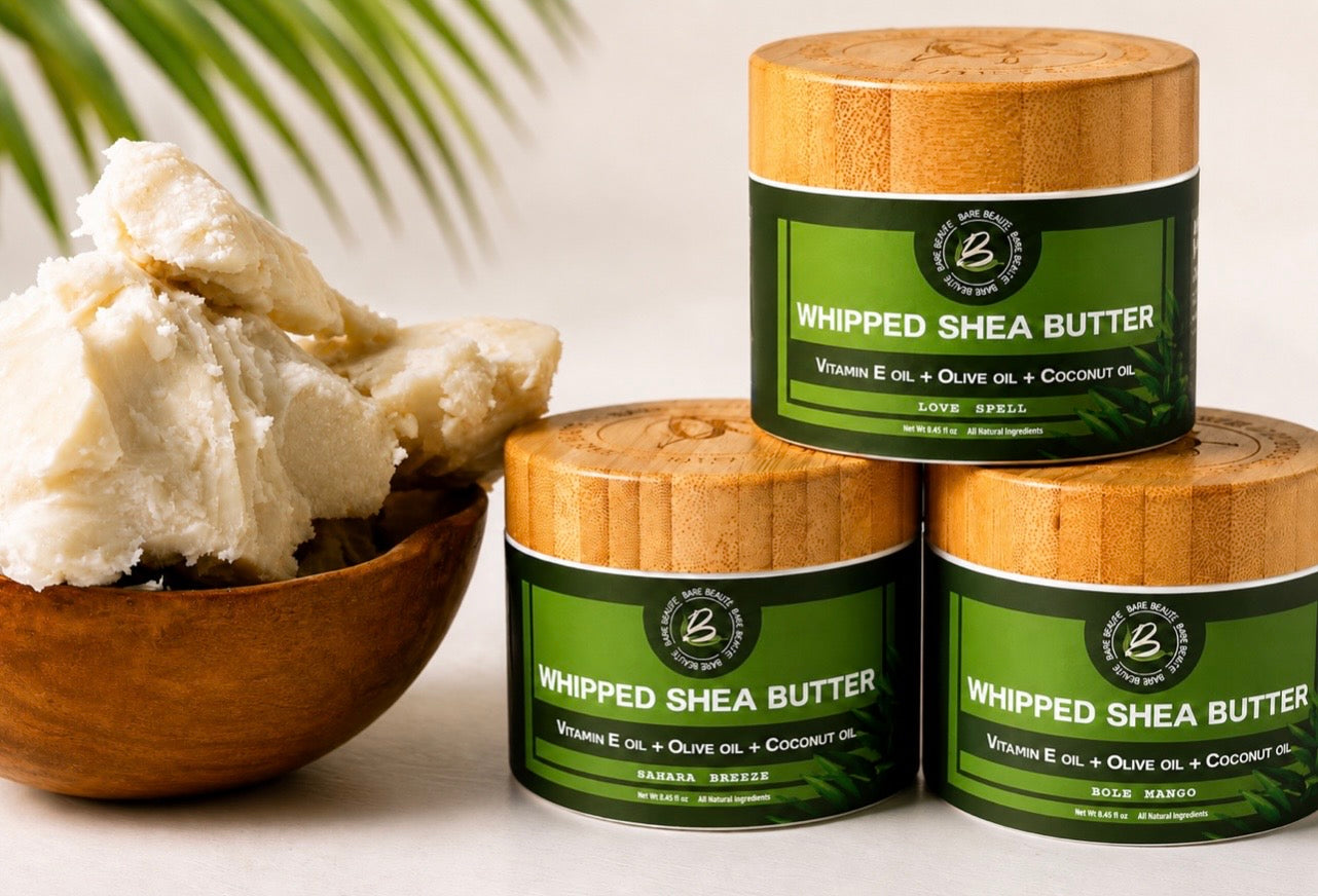 Three jars of Whipped Shea Butter with bamboo lids and green labels are arranged on a light surface, alongside a wooden bowl filled with pale whipped shea butter. A blurred palm leaf appears in the background.
