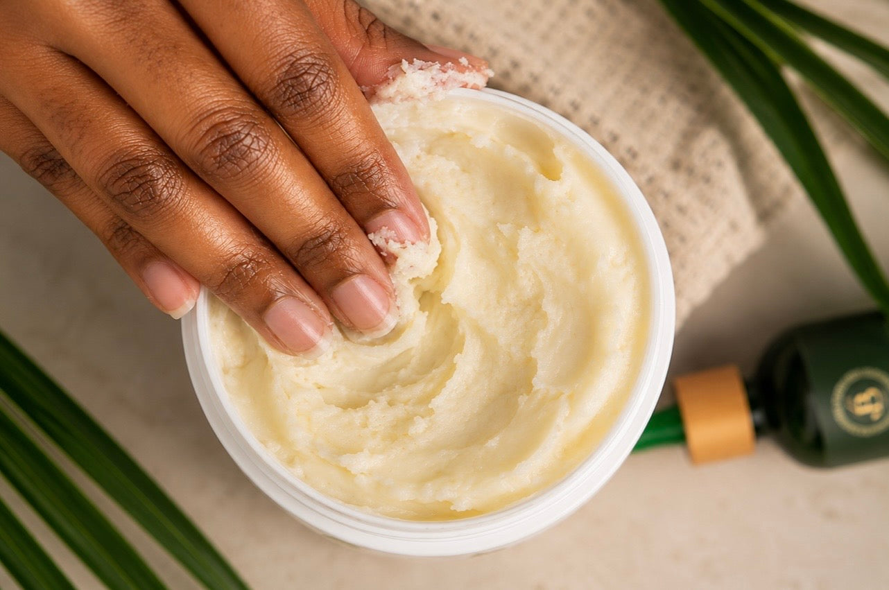 A close-up of a hand applying a thick, creamy off-white body scrub from an open white jar, with a dark green bottle and green leaves in the background.