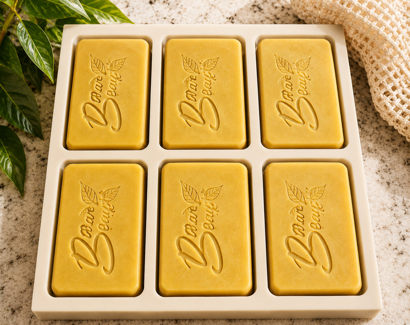 Six yellow bars of Bare Beaute Essentials Turmeric and Shea Butter soapz arranged in a white tray on a speckled stone surface, with green leaves and a beige cloth partially visible around the edges.