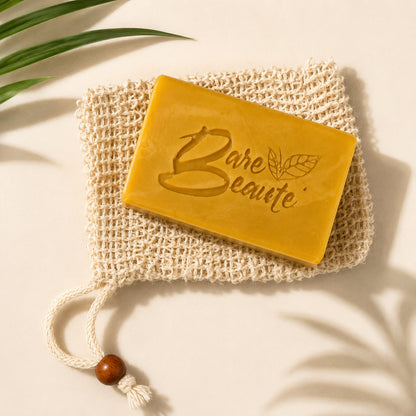A rectangular yellow bar of Bare Beaute Turmeric and Shea Butter soap resting on a cream-colored woven mesh soap bag with a drawstring and wooden bead, styled with green leaves on a warm beige background.